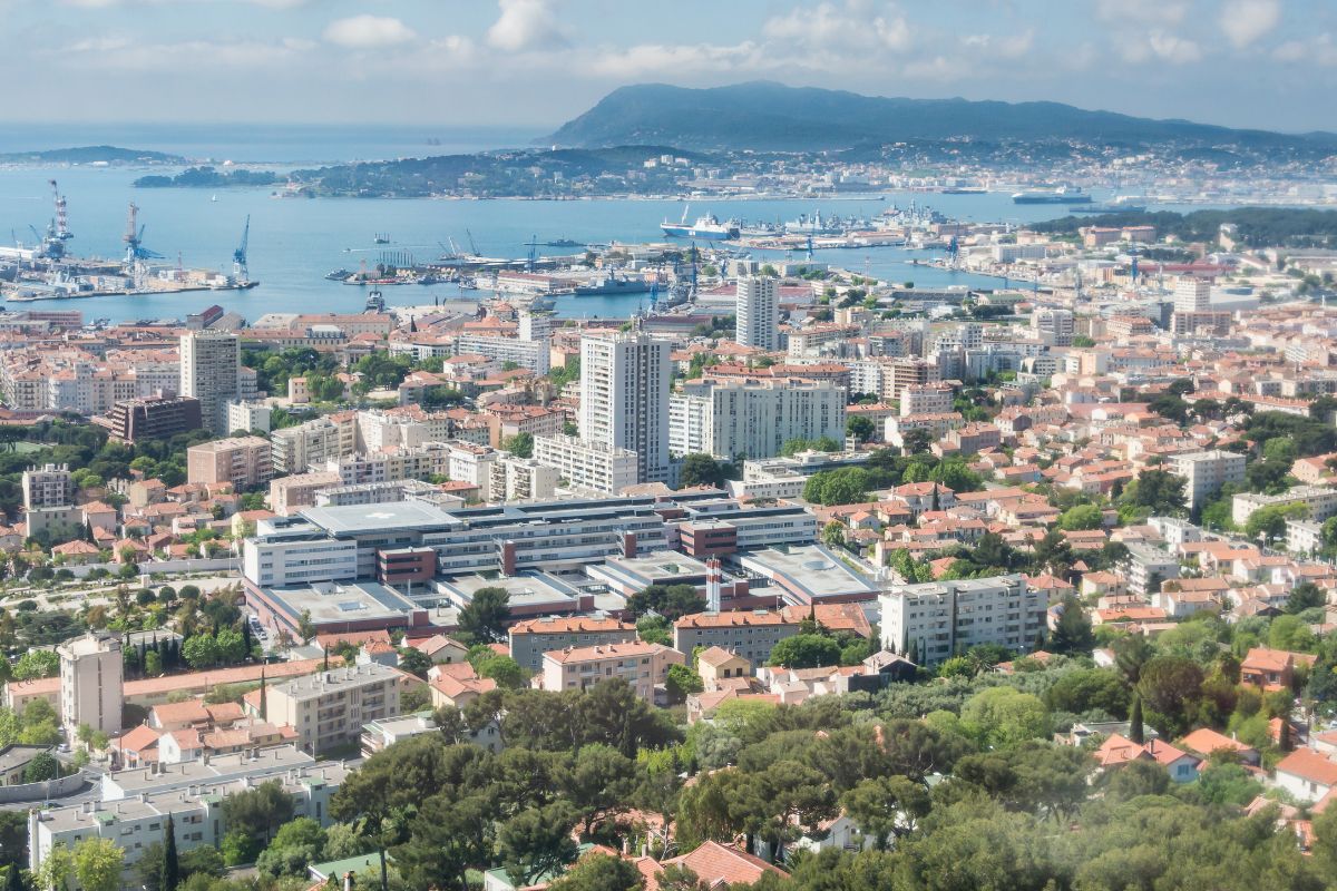Vue aérienne d'une ville côtière avec de nombreux bâtiments, des espaces verts et plusieurs navires amarrés dans un port, sur fond de montagnes et d'un ciel partiellement nuageux.