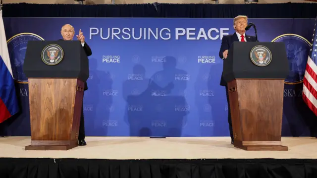 Poutine, s'exprimant depuis un podium et pointant du doigt de la main gauche, tandis que Trump, debout à un autre podium à la gauche de Poutine, regarde droit devant lui, à Anchorage, en Alaska, le 15 août 2025. Un panneau derrière eux porte l'inscription "Poursuivre la paix".