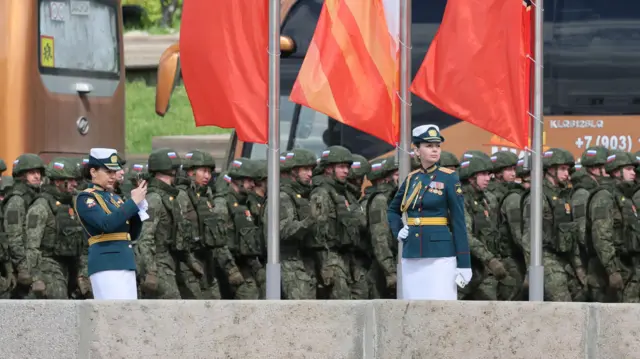 Le 7 mai 2025, deux militaires russes, vêtues de tuniques vertes ornées de médailles, prennent des photos tandis que passent des soldats en treillis verts. Des drapeaux rouges et oranges flottent au-dessus du groupe.