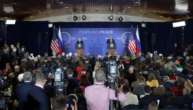 Des journalistes font face à une estrade où Poutine et Trump se tiennent à des pupitres. Au premier plan, deux caméras sur trépieds sont manipulées par deux hommes. Les autres journalistes sont assis, serrés les uns contre les autres.