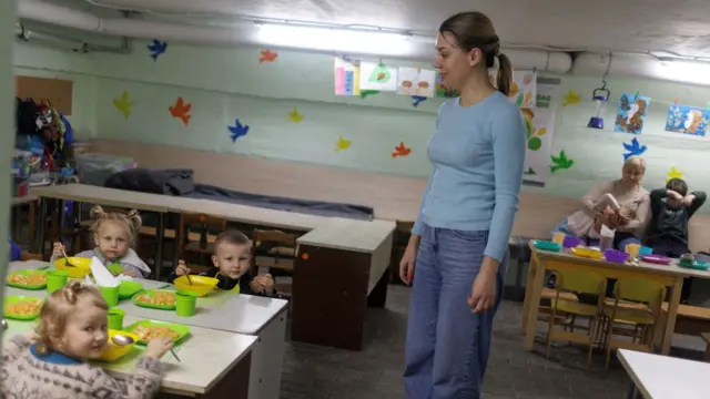 Maryna Ponomarenko, 40 ans, employée d'une école maternelle, sourit en observant des enfants attablés dans des bols, le 10 octobre 2025. Ils se trouvent dans un abri anti-bombes et la tête de Mme Ponomarenko touche presque le plafond. Des découpages d'oiseaux multicolores sont collés sur les murs vert clair. Une femme est assise à l'arrière-plan, une fillette penchée sur ses genoux et un autre enfant, assis à sa gauche, tirant sur le cordon de son pantalon. Des dessins sont accrochés à des pinces fixées à des tuyaux.