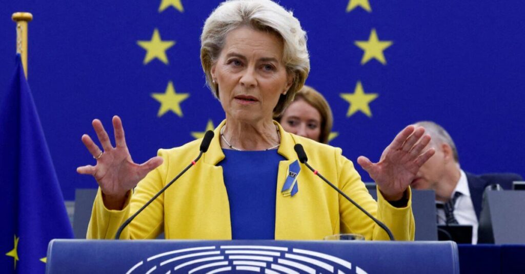 Femme s’exprimant à la tribune du Parlement européen, vêtue aux couleurs de l’UE, avec des drapeaux européens en arrière-plan.