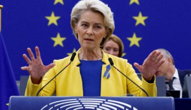 Femme s’exprimant à la tribune du Parlement européen, vêtue aux couleurs de l’UE, avec des drapeaux européens en arrière-plan.