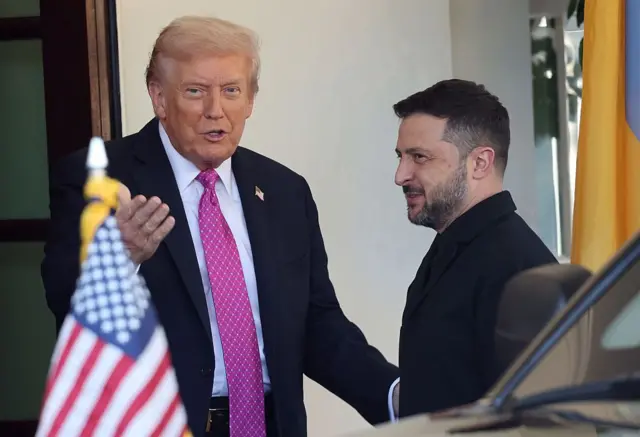 Trump salue Zelensky à l'entrée de la Maison-Blanche et lui adresse quelques mots sur un ton apparemment léger. Zelensky sourit. Au premier plan, le drapeau américain flotte à l'avant d'une voiture.