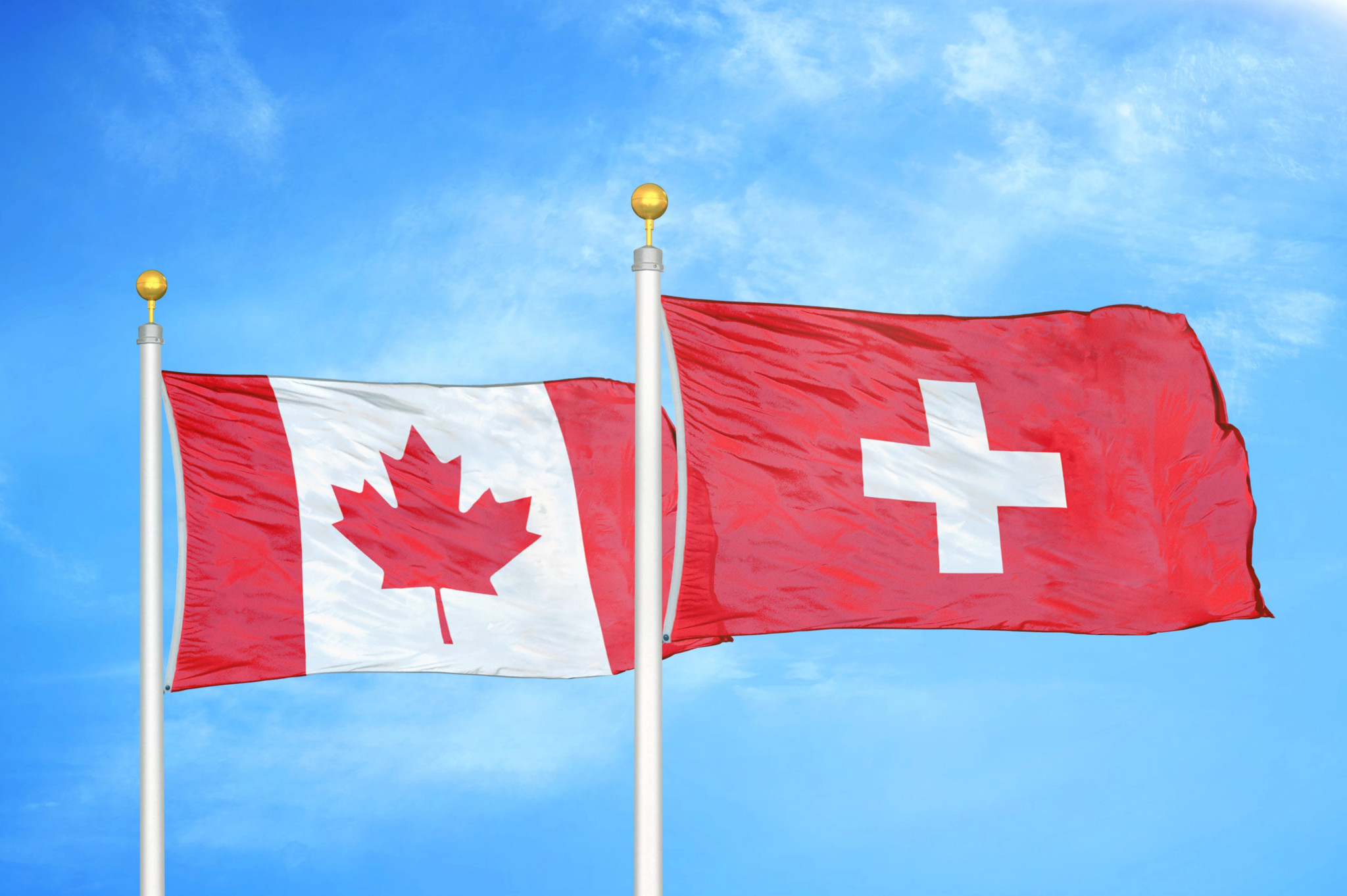Les drapeaux du Canada et de la Suisse sur des mâts avec un ciel bleu nuageux en arrière-plan.