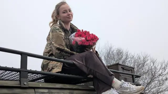 Une fille en veste militaire avec des fleurs est assise sur une voiture.