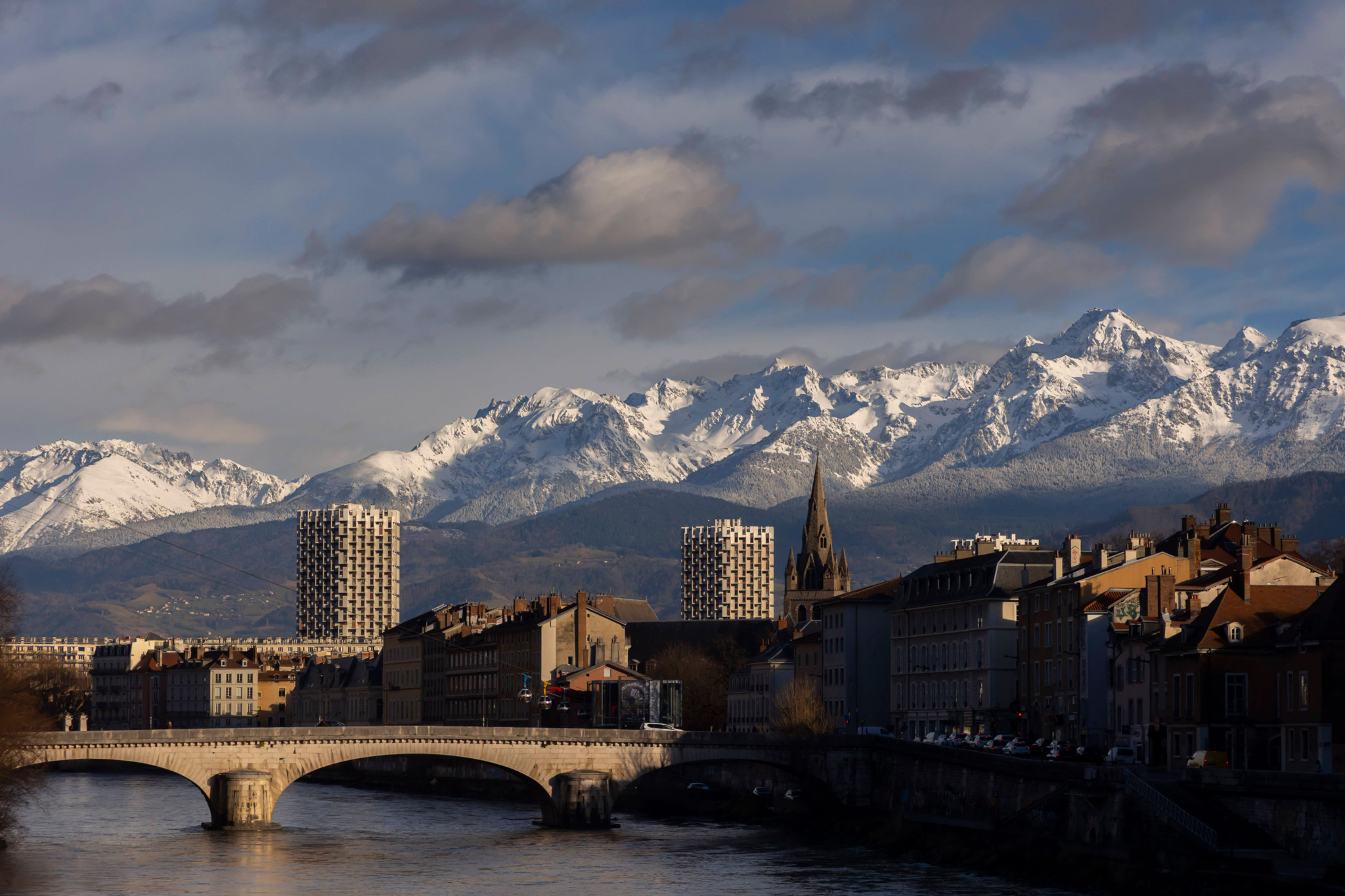 Vue panoramique de Grenoble avec montagnes enneigées des Alpes et rivière Isère sous un ciel nuageux.