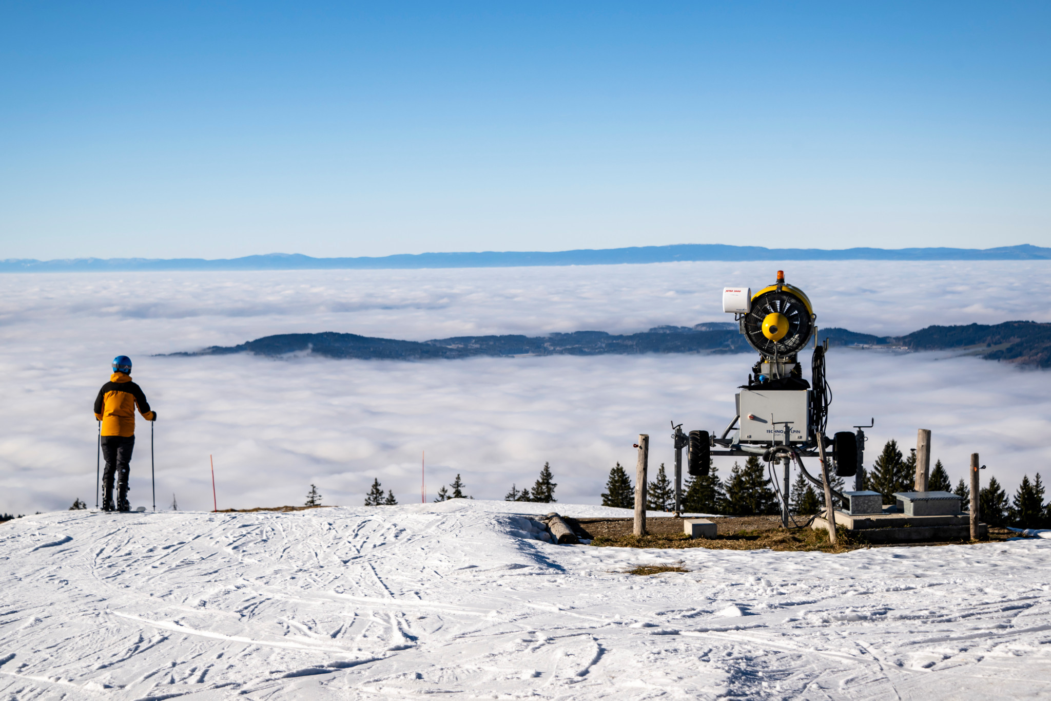 Des skieurs sur les pistes enneigées de La Berra avec un canon à neige en avant-plan et une mer de brouillard à l’horizon, dans les Préalpes fribourgeoises.