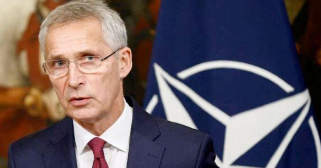 Homme en costume sombre et cravate rouge prenant la parole devant un drapeau de l’OTAN, expression sérieuse dans un contexte diplomatique ou sécuritaire.