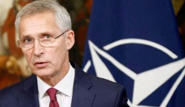 Homme en costume sombre et cravate rouge prenant la parole devant un drapeau de l’OTAN, expression sérieuse dans un contexte diplomatique ou sécuritaire.