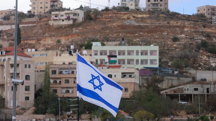 Un drapeau palestinien est visible sur un bâtiment situé en face d’un drapeau national israélien placé à l’entrée du village d’As-Sawiyah, en Cisjordanie occupée, le 16 novembre 2025, après que des colons aient placé des centaines de drapeaux israéliens sur les routes publiques et à l’entrée des villages palestiniens.