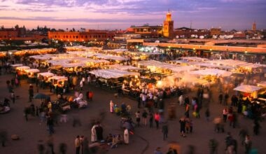 Marché en plein air bondé au coucher du soleil avec de nombreux étals couverts par des auvents blancs, des gens qui marchent et des bâtiments avec une mosquée en arrière-plan sous un ciel violet.