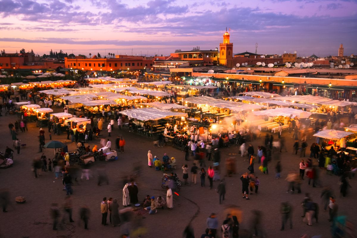 Marché en plein air bondé au coucher du soleil avec de nombreux étals couverts par des auvents blancs, des gens qui marchent et des bâtiments avec une mosquée en arrière-plan sous un ciel violet.