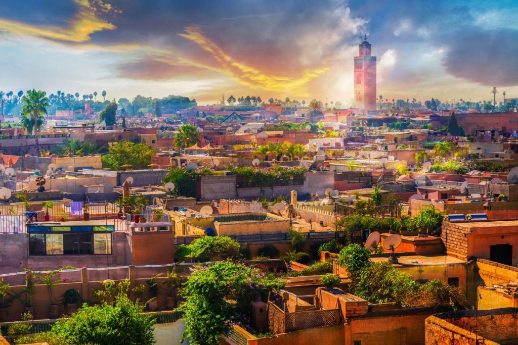 Paysage urbain de Marrakech au coucher du soleil, montrant des toits plats, des antennes paraboliques, une végétation luxuriante et la tour de la mosquée Koutoubia sous un ciel spectaculaire.