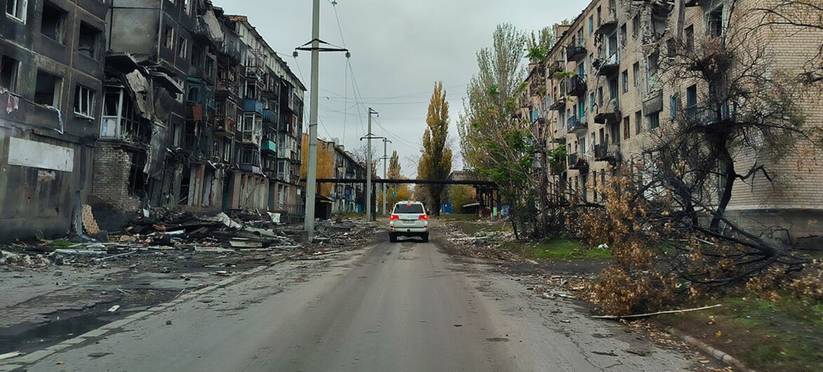 Une voiture blanche conduit dans une rue endommagée en Ukraine, entourée de bâtiments d'appartements fortement bombardés avec des fenêtres cassées et des façades charbonnées.