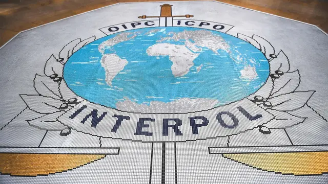 Une mosaïque au sol du bâtiment d'Interpol représentant le logo de l'organisation. Elle comporte une carte du monde dans un cercle, avec les mers représentées en bleu et les terres en gris. Derrière la carte se trouve une épée, et en dessous, une balance en or et le mot « Interpol ».