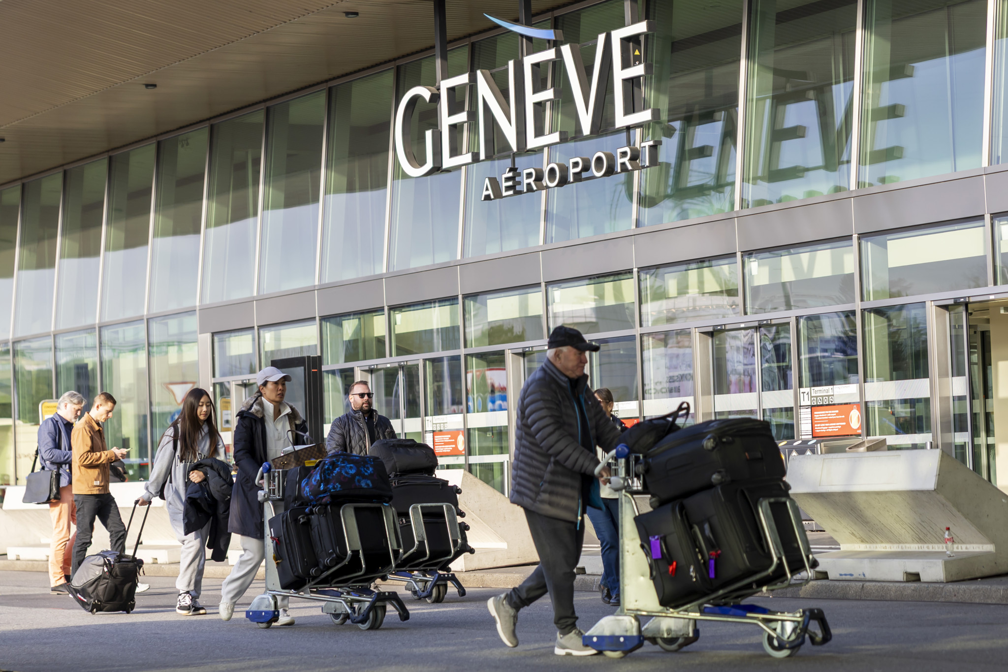 Des passagers avec des chariots à bagages arrivent devant le hall du terminal 1 de Genève Aéroport.