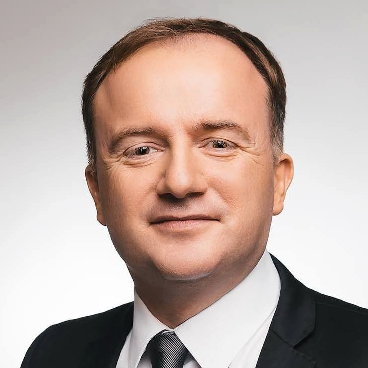 Portrait d’un homme en costume avec une cravate, souriant légèrement, sur un fond uni.