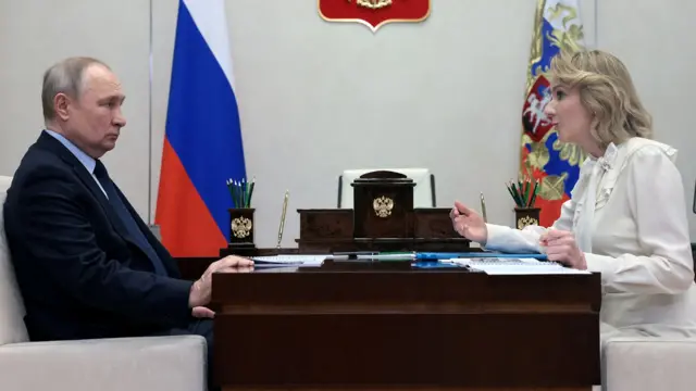 Vladimir Poutine et Maria Lvova-Belova sont assis de part et d'autre d'un bureau. Il est à gauche et la regarde pendant qu'elle parle. Il porte un costume sombre et une cravate, elle porte une robe blanche. Un drapeau russe est visible en arrière-plan. 