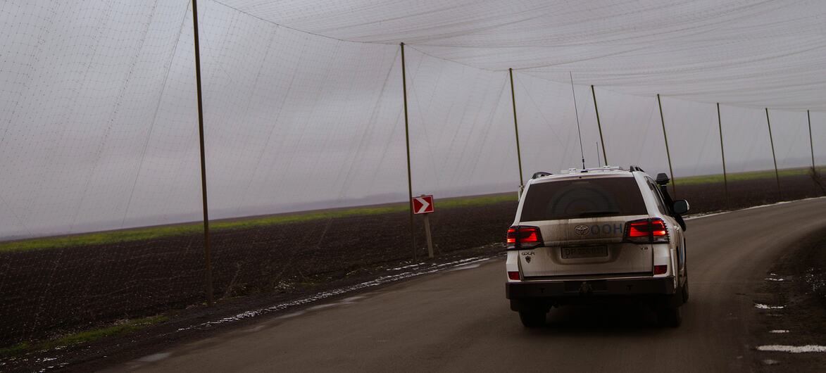 Un SUV blanc conduit le long d'une route rurale en Ukraine, passant sous un grand filet agricole soutenu par des poteaux.