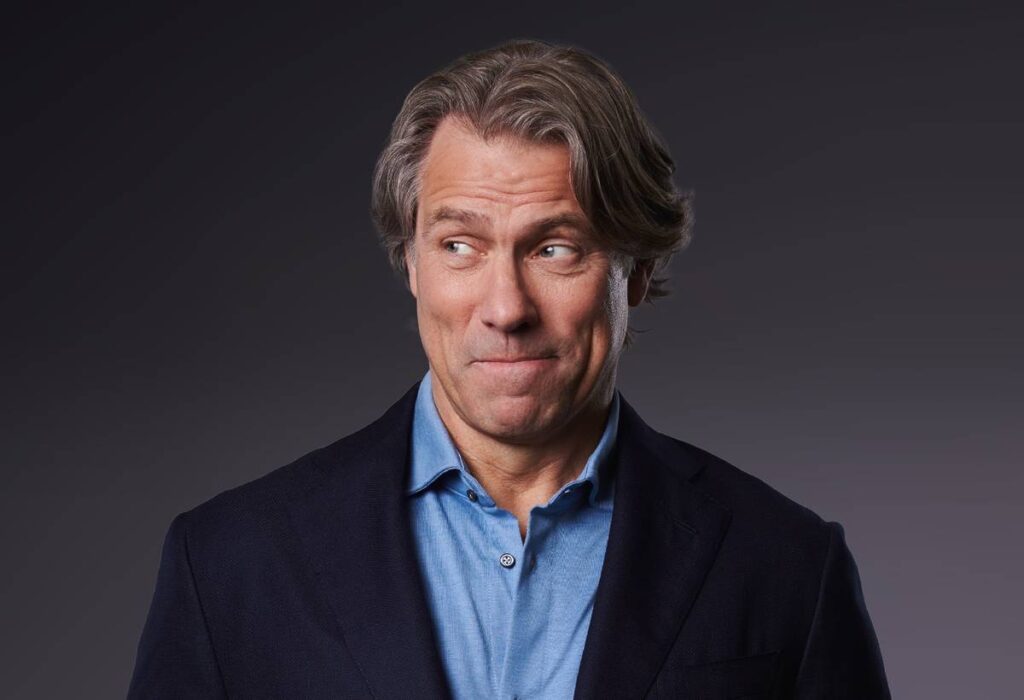 L'humoriste John Bishop sourit et regarde hors champ, sur le côté.