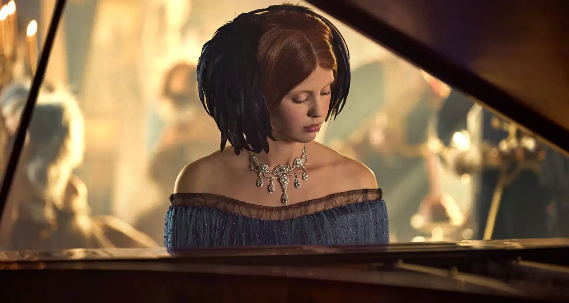 Woman in elegant gown and necklace plays piano at a dimly lit formal event with people in the background.