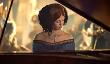 Woman in elegant gown and necklace plays piano at a dimly lit formal event with people in the background.