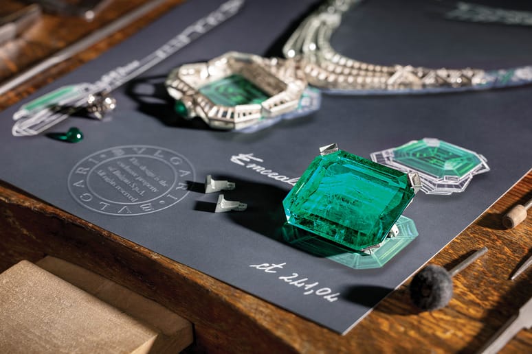 Emerald gemstones and jewelry designs displayed on a table with drawing tools and sketches.