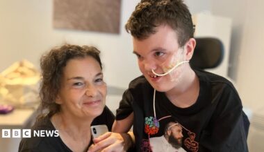 Sam, is a woman with brown wavy hair. She is wearing a black top, kneeling next to Alex's wheelchair smiling for the camera. Alex is a boy in a black T-shirt, with a feeding tube attached to his nose.