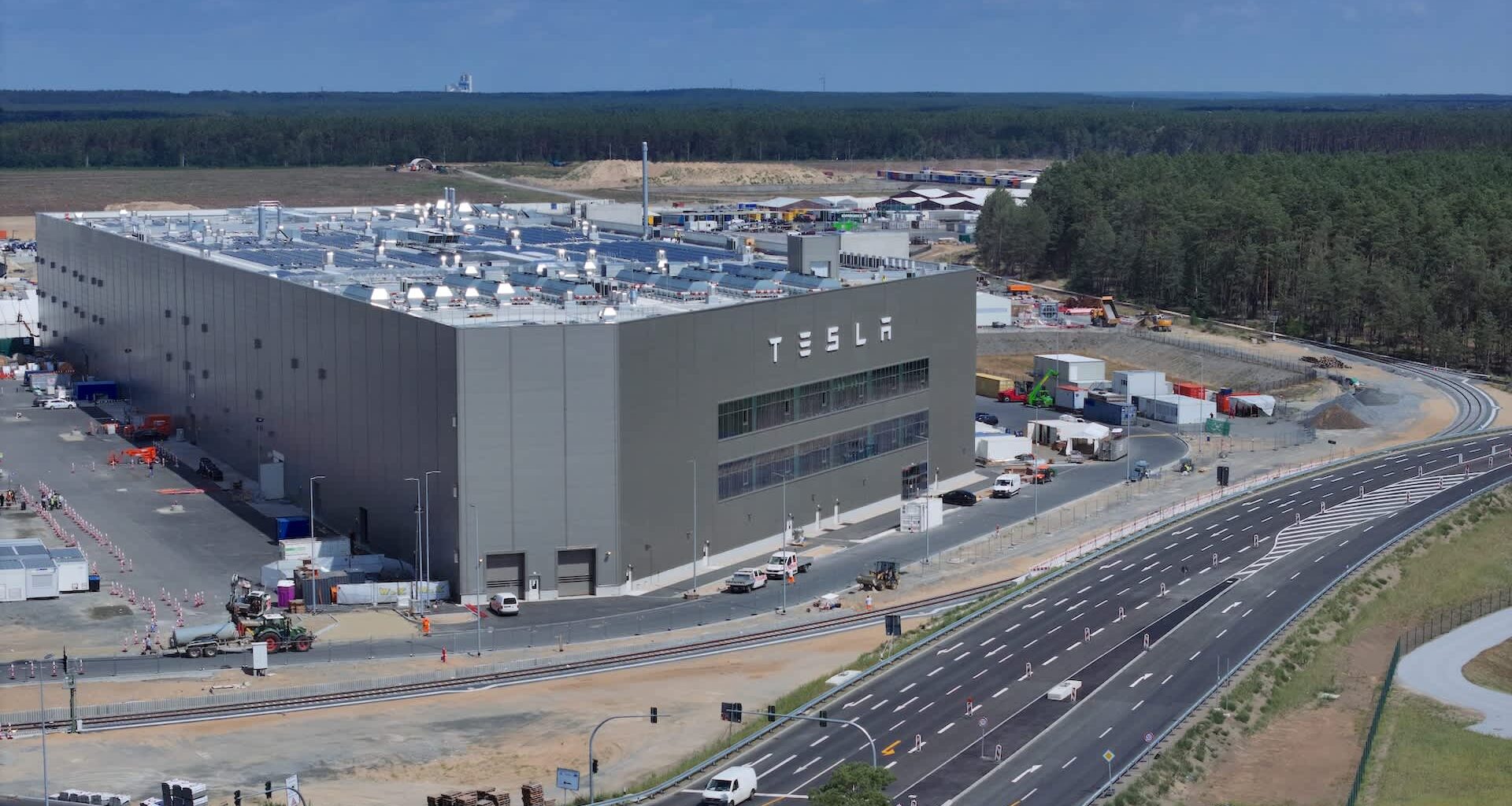 A large Tesla factory building in Berlin with construction activity and vehicles in the surrounding area on a sunny day.