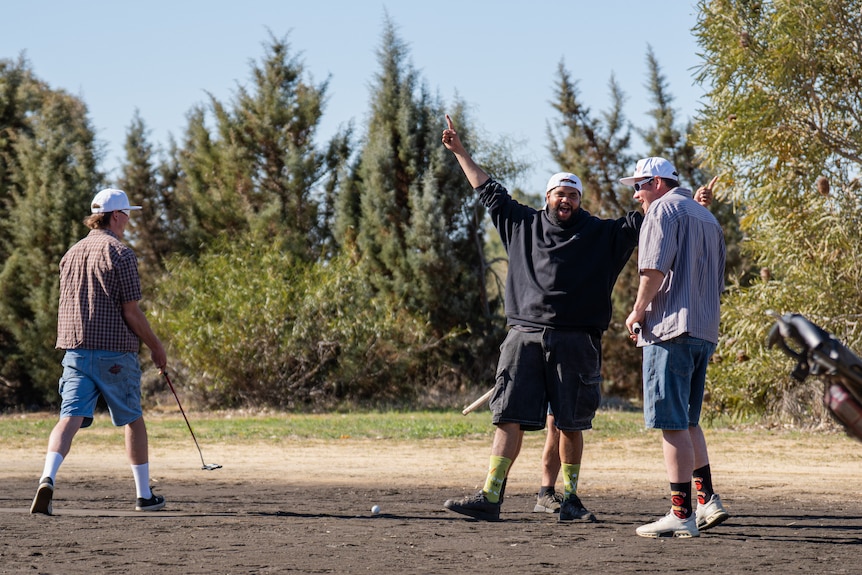 Three young men on a golf course, one has hands in the air celebrating