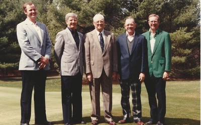 Five people in jackets and dress pants stand side by side on a golf course, with tall evergreen trees in the background. The vintage photograph shows the group posing outdoors on a sunny day.