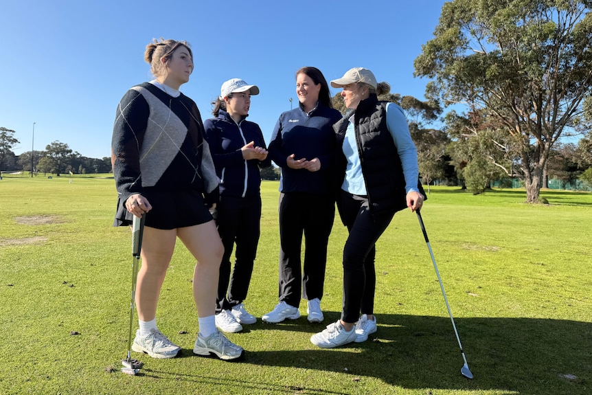 Four women stand together on a golf course. Two are wearing caps and two hold golf clubs.WEo
