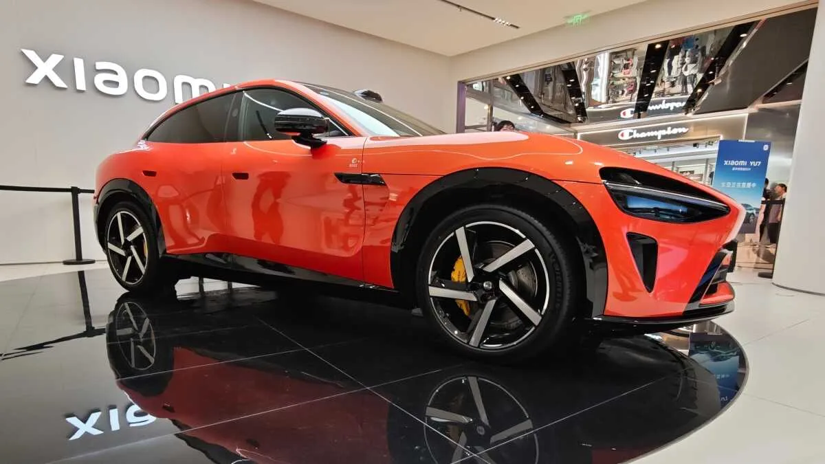 Red sports car on a showroom platform with glossy black floor and a Xiaomi sign in the background, showcasing a sleek aerodynamic design and large alloy wheels.