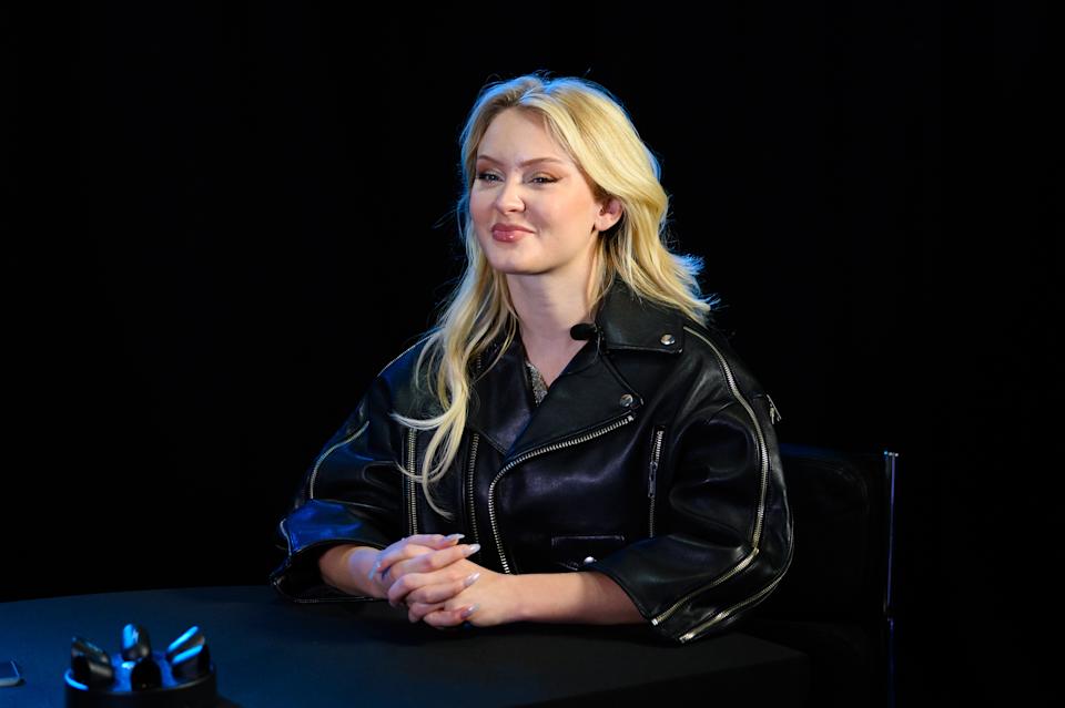 Zara Larsson with long hair in a leather jacket sitting at a table against a background