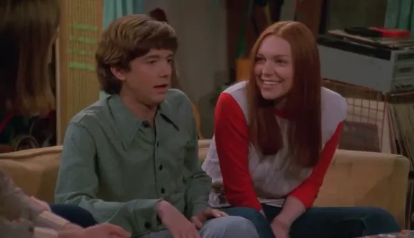 Two seated people smiling and talking in a casual setting, one wearing a button-up shirt, the other a raglan sleeve shirt