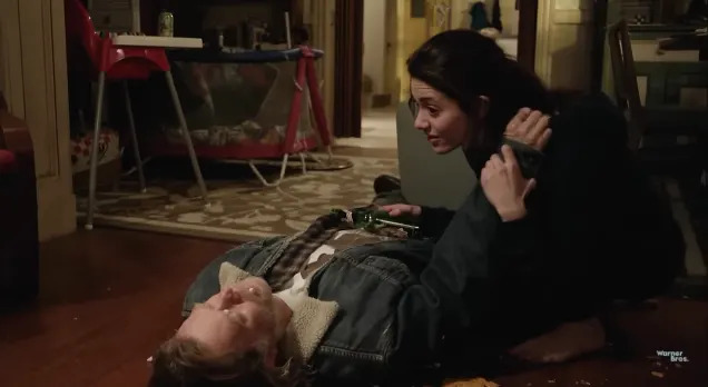 A woman kneels beside a man lying on a messy living room floor, holding a bottle. The room is cluttered with objects and furniture