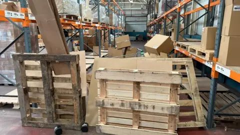 Jordan Dales Several wooden pallets are propped up against each other in a warehouse next to an aisle of empty goods other than cardboard boxes.