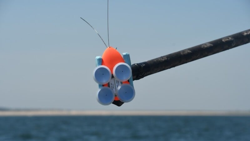 A small, orange and blue floating device with antennas is attached to a pole above a body of water, with a distant shoreline visible in the background under a clear sky.