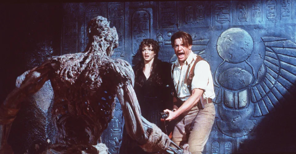 Scene from a movie with two actors facing a mummy in an Egyptian setting. The man holds a weapon, and the woman looks shocked