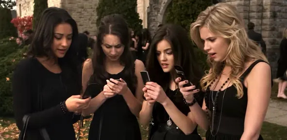 Four people in stylish black outfits focus intently on their phones in an outdoor setting.