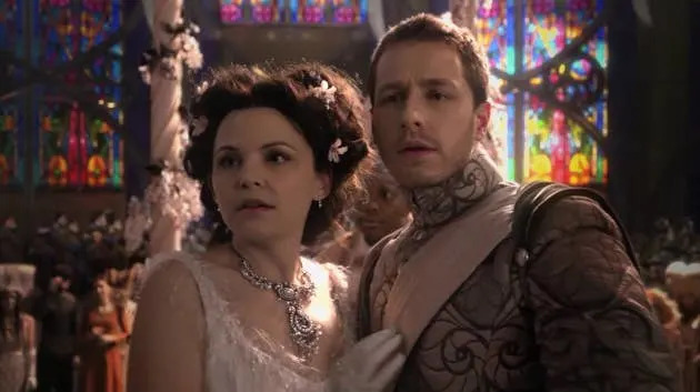 Two people dressed in elaborate, fantasy-style costumes stand together in a setting with stained glass windows, looking concerned