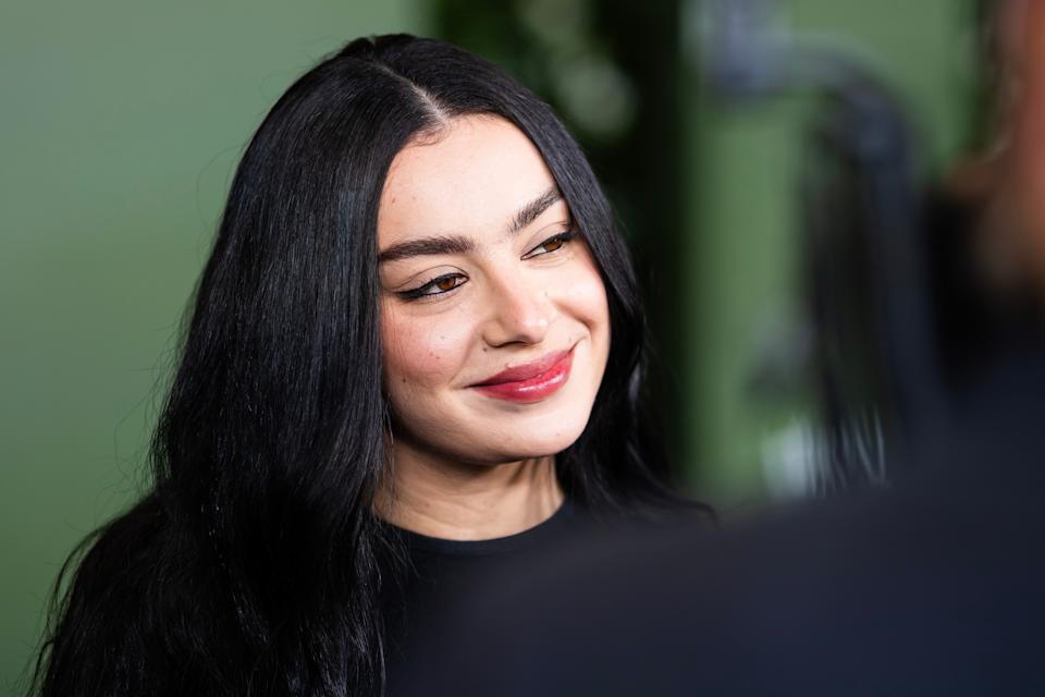 Charli XCX with long dark hair is smiling, wearing a simple, elegant outfit. The setting appears to be a casual event