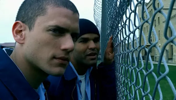 Two people in prison uniforms stand by a chain-link fence, looking thoughtful and serious, with a prison building in the background