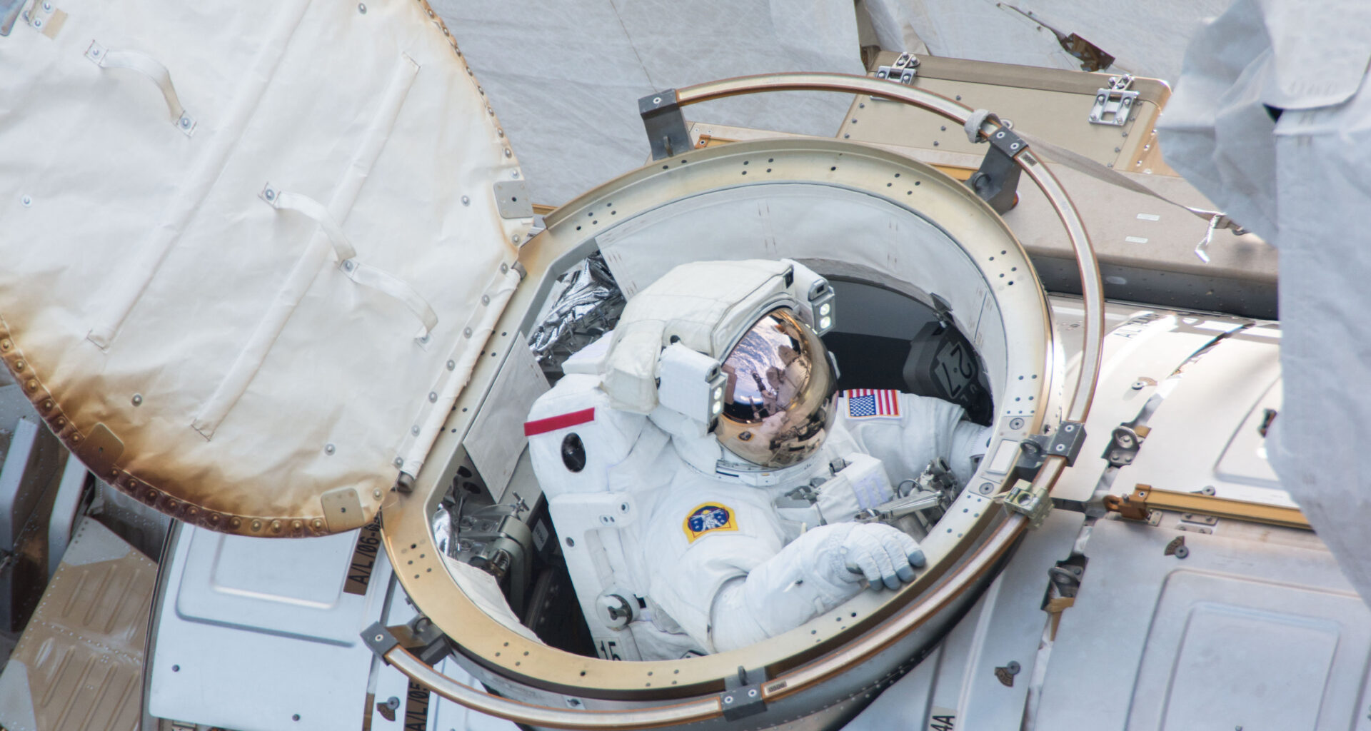 An astronaut in a white spacesuit comes out of a hatch on the International Space Station. We can see parts of the space station in the photo.