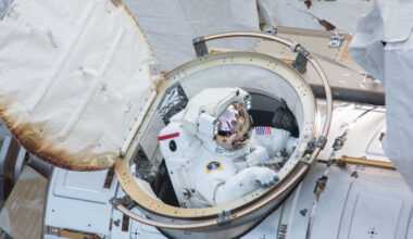 An astronaut in a white spacesuit comes out of a hatch on the International Space Station. We can see parts of the space station in the photo.