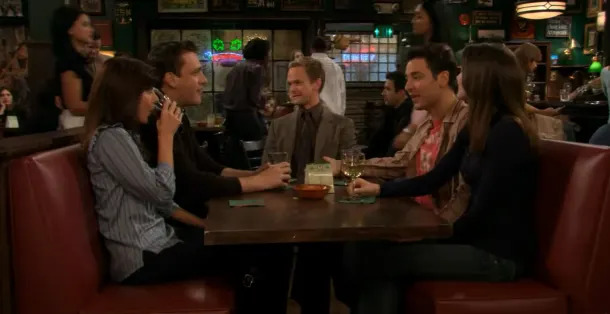 A group of five people are sitting in a booth at a lively bar, engaging in friendly conversation