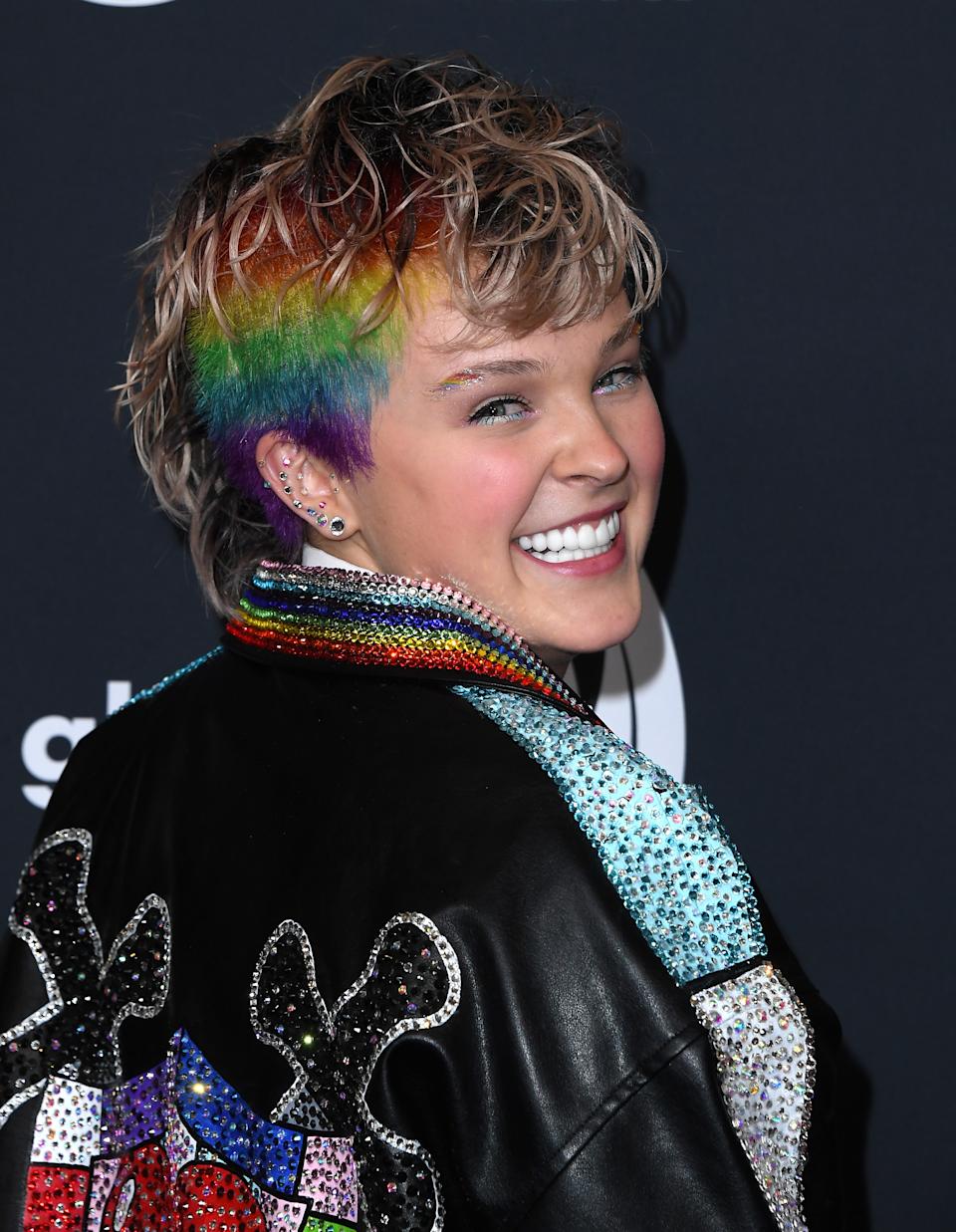 JoJo Siwa smiling on the red carpet, wearing a brightly decorated jacket adorned with sequins and rainbow highlights in their short hair