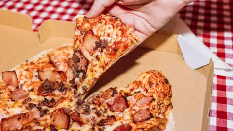 Getty Images A hand picking up a slide of ham pizza from a takeaway box on a red and white gingham tablecloth.