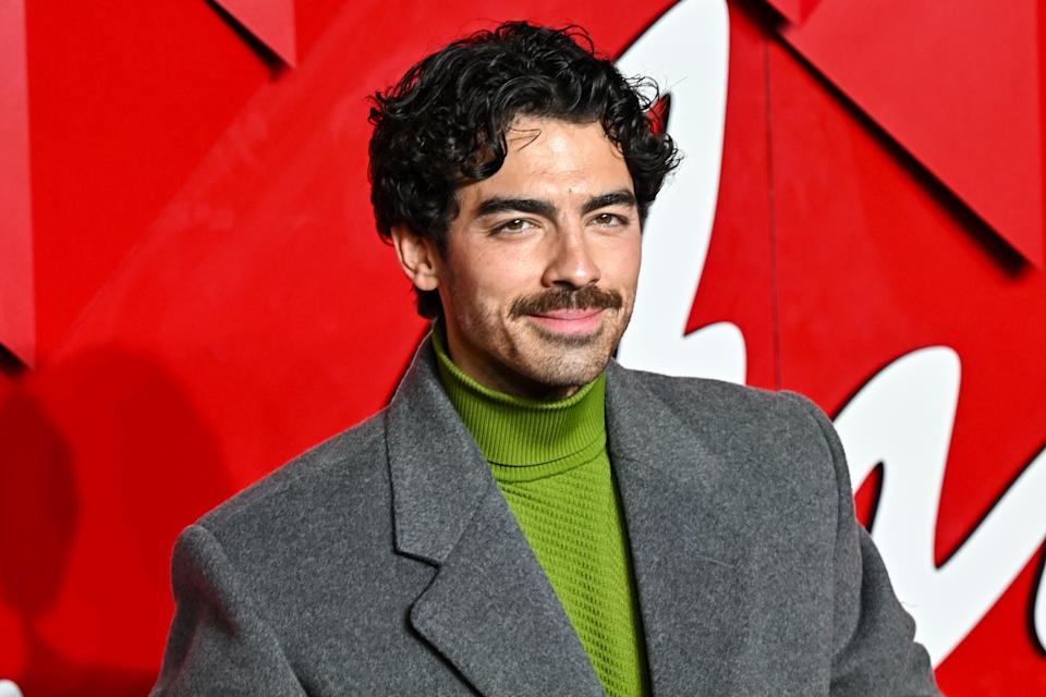 Joe Jonas in a coat and turtleneck on a red carpet with a textured backdrop
